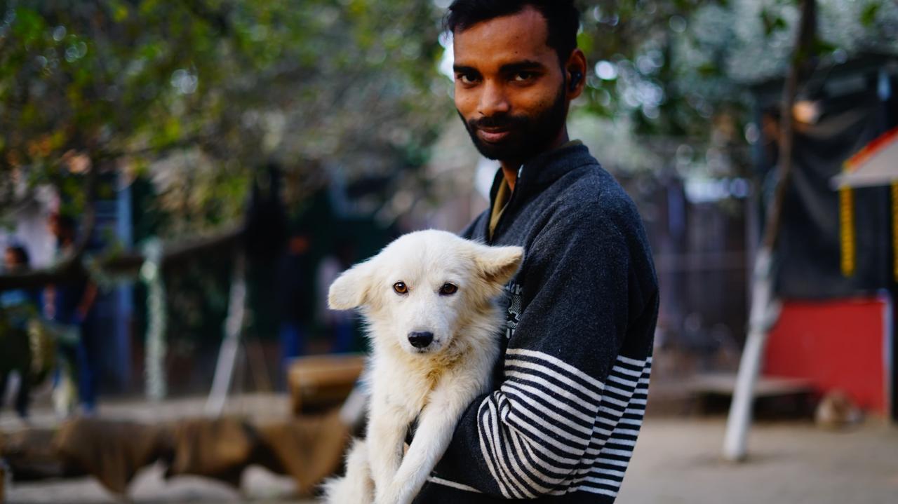 A man holding a small white dog in his arms, with trees and blurred buildings in the background.